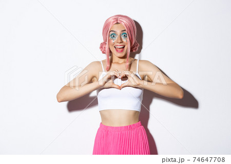 Image of excited smiling girl like something, showing heart gesture and looking amazed, wearing halloween costume with bright makeup and pink wig 74647708