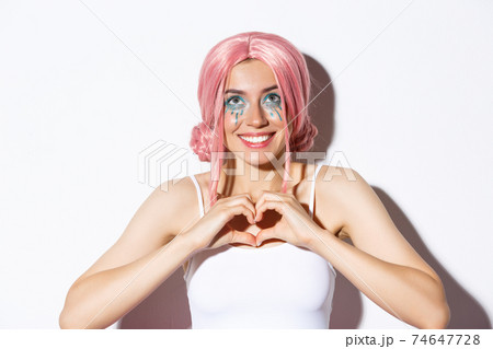 Close-up of hopeful smiling girl in pink party wig, looking up and showing heart sign, standing over white background 74647728