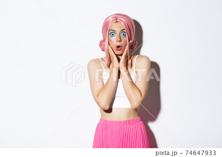 Portrait of surprised caucasian girl in pink wig and bright makeup, gasping amazed and stare at camera in awe, standing over white background in halloween party costume 74647933