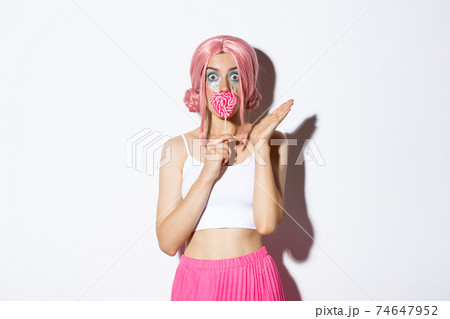 Image of flirty female model with pink wig and bright makeup, kissing heart-shaped candy and looking surprised, celebrating halloween Image of flirty female model with pink wig and bright makeup, kissing heart-shaped candy and looking surprised, celebrating halloween 74647952