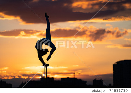 Flexible female circus Artist keep balance and doing contortion on the rooftop against dramatic sunset and cityscape. Motivation, passion and achievement concept  74651750