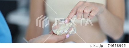 Woman patient with elastic bandage on her chest touches a silicone implant in doctor's hand close-up Woman patient with elastic bandage on her chest touches a silicone implant in doctor's hand close-up 74658946