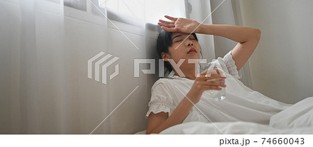 An ill woman drinks water and consuming a pill while lying on the bed in the bedroom. An ill woman drinks water and consuming a pill while lying on the bed in the bedroom. 74660043