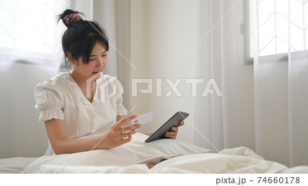 Asian woman is shopping online with a computer tablet while sitting on the bed in a comfortable bedroom. Asian woman is shopping online with a computer tablet while sitting on the bed in a comfortable bedroom. 74660178