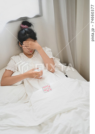 An ill woman holds a glass of water and consuming a pill while lying on the bed in the bedroom. 74660377