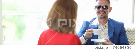 Happy man in blue jacket and sunglasses hold white mug and saucer in his hand and look at woman. Happy man in blue jacket and sunglasses hold white mug and saucer in his hand and look at woman. 74664350