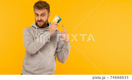 Attractive man in a gray hoodie points a finger at the credit card on a yellow background Attractive man in a gray hoodie points a finger at the credit card on a yellow background 74666635