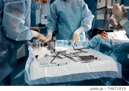 close up surgeons hand taking scissors, forceps and surgical instruments on table for operation with colleagues performing work in operation room at hospital, emergency case, surgery, medical concept 74671919