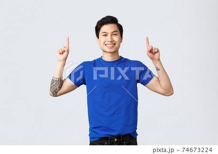 Different emotions, people lifestyle and advertising concept. Cheerful handsome asian man in blue t-shirt pointing fingers up. Guy advertising and inform about new promo, smiling camera Different emotions, people lifestyle and advertising concept. Cheerful handsome asian man in blue t-shirt pointing fingers up. Guy advertising and inform about new promo, smiling camera 74673224
