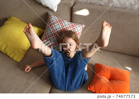 a little beautiful girl in a blue jumpsuit is lying at home on the sofa with her legs raised up. a little beautiful girl in a blue jumpsuit is lying at home on the sofa with her legs raised up. 74677573
