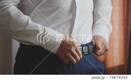 A smart man puts a belt on his pants with a white shirt to keep the pants on his hips. Getting ready early in the morning for work in a room at home. Close-up 74679265