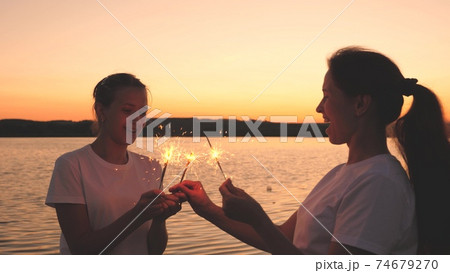 Happy girls set fire to sparklers at sunset in the sky and laugh. Young people greet the sunrise with beautiful lights. Girlfriends travel. Rest during the holidays. Celebrating your birthday with fun 74679270
