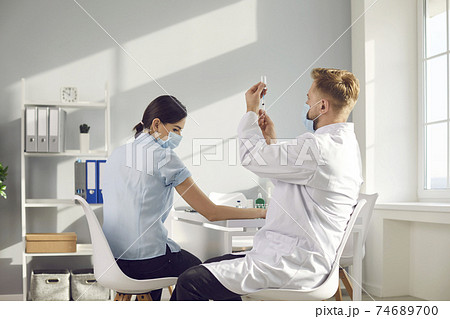 Male nurse drawing antiviral vaccine out of vial before giving shot to young woman 74689700