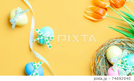 Easter frame. Colorful egg with tape ribbon, spring tulips, feathers on pastel yellow background in Happy Easter decoration. Festive design in flat lay. 74692040