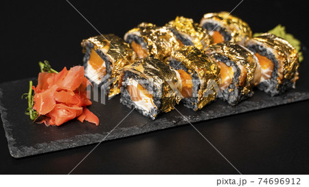 Custom sushi roll with nori, cuttlefish ink, fresh salmon, cream cheese, pepper pumpkin, eel, unagi sauce and edible gold leaf with red ginger, wasabi on a square plate on black table and background. 74696912
