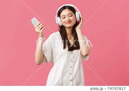 Beauty, people emotions and technology concept. Carefree and pretty asian girl enjoying music in wireless headphones, close eyes and smiling listening favorite song, holding smartphone and dancing 74707817