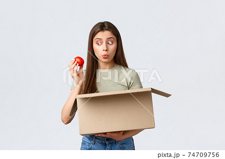 Online home delivery, internet orders and grocery shopping concept. Excited cute female customer receive groceries from internet store, take-out tomato and smiling amused 74709756