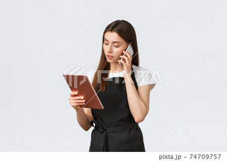 Grocery store employees, small business and coffee shops concept. Cute smiling saleswoman in black apron confirm order, talking to customer on phone and looking at digital tablet 74709757