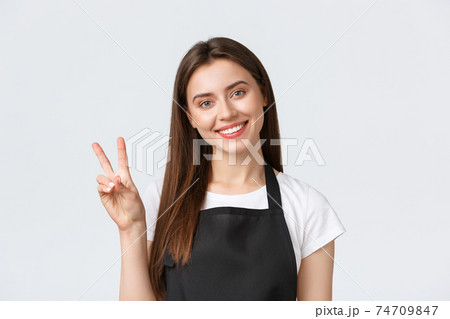 Employees, job employment, small business and coffee shop concept. Close-up of friendly-looking smiling female barista, cafe worker in black apron greeting guests and show peace sign Employees, job employment, small business and coffee shop concept. Close-up of friendly-looking smiling female barista, cafe worker in black apron greeting guests and show peace sign 74709847