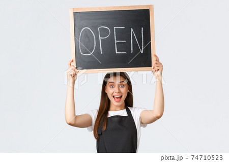 Grocery store employees, small business and coffee shops concept. Cheerful happy barista in black apron raise open sign, inform customers that shop or cafe working after covid pandemic Grocery store employees, small business and coffee shops concept. Cheerful happy barista in black apron raise open sign, inform customers that shop or cafe working after covid pandemic 74710523
