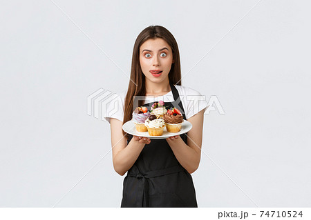 Grocery store employees, small business and coffee shops concept. Cute young female barista in black apron holding tasty dessert, lick lips desire have bite of delicious cupcakes, white background Grocery store employees, small business and coffee shops concept. Cute young female barista in black apron holding tasty dessert, lick lips desire have bite of delicious cupcakes, white background 74710524