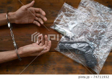Handcuffed hands of criminal suspect on wooden table and handgun with jackknife in transparent plastic packs as a crime scene evidence for investigation 74711932
