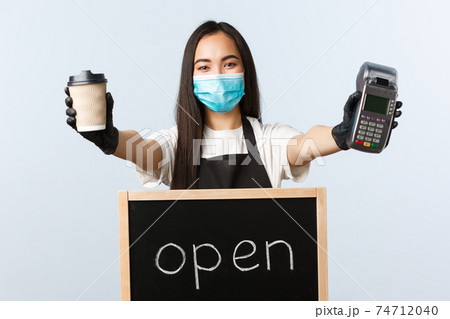 Small business, covid-19 pandemic, preventing virus and contactless orders concept. Smiling nice asian barista, female cafe worker with open sign, giving takeaway coffee and POS terminal 74712040