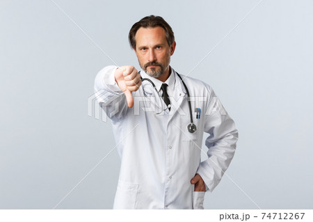 Covid-19, coronavirus outbreak, healthcare workers and pandemic concept. Disappointed serious male doctor in white coat, frowning displeased and show thumb-down, disapprove action 74712267