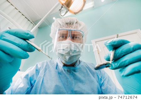 a dentist wearing a protective mask sits nearby and holds instruments in his hands before treating a patient in the dental office a dentist wearing a protective mask sits nearby and holds instruments in his hands before treating a patient in the dental office 74712674