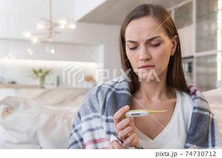 Woman sits on the sofa and looks at the thermometer Woman sits on the sofa and looks at the thermometer 74730712