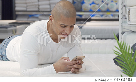 Young African man using smart phone while trying new bed at furniture store 74733482
