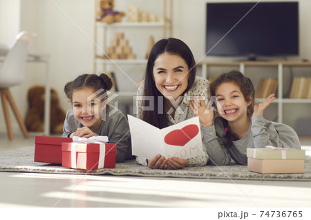 Mother with a postcard in her hands lying on the floor with her daughters among the gifts. Mother with a postcard in her hands lying on the floor with her daughters among the gifts. 74736765