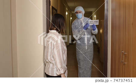 Woman opening door at home for doctor in PPE medical biohazard suit, vaccination against coronavirus 74737451