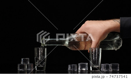 Bartender pouring up vodka from bottle into two shots glasses with ice cubes on black background Bartender pouring up vodka from bottle into two shots glasses with ice cubes on black background 74737541