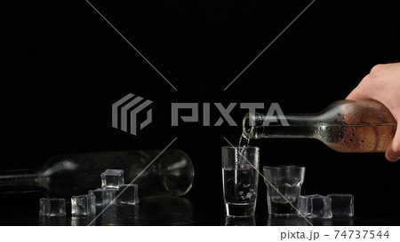 Bartender pouring up two shots of vodka with ice cubes from bottle into glasses on black background 74737544