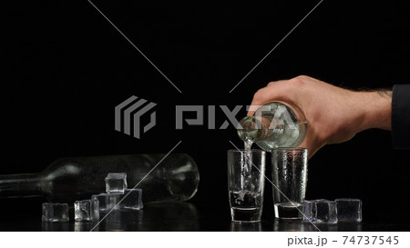 Bartender pouring up vodka from bottle into two shots glasses with ice cubes on black background 74737545