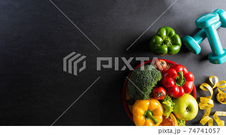 Top view of healthy food in plate with dumbbells and measure tape for cholesterol diet control on dark background. World health day and medical concept. 74741057