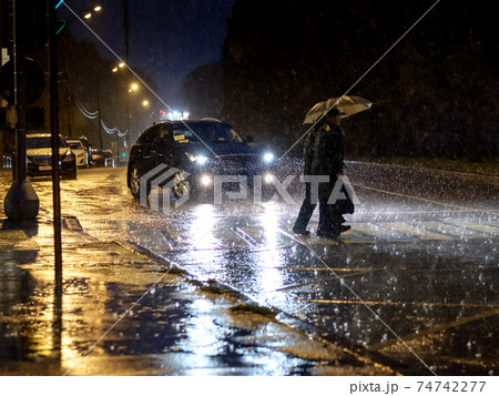 View of a pedestrian crossing in the city at night during a heavy downpour. Silhouettes of people with umbrellas. 74742277