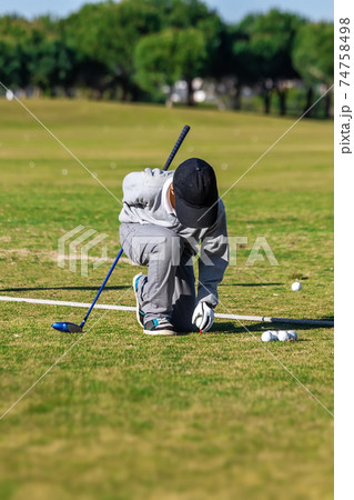 Golfer boy crouching down and placing a ball on a tee support, on a grass course. Preparing his next shot. Golfer boy crouching down and placing a ball on a tee support, on a grass course. Preparing his next shot. 74758498