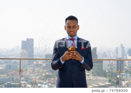 Portrait of handsome young African businessman wearing suit while smiling and using mobile phone 74768930