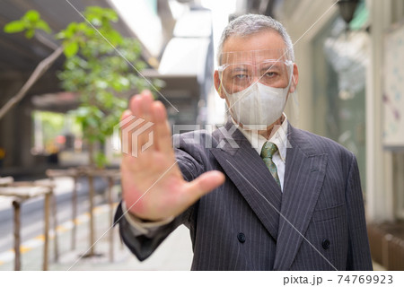 Stressed mature Japanese businessman with mask and face shield showing stop gesture in the city Stressed mature Japanese businessman with mask and face shield showing stop gesture in the city 74769923