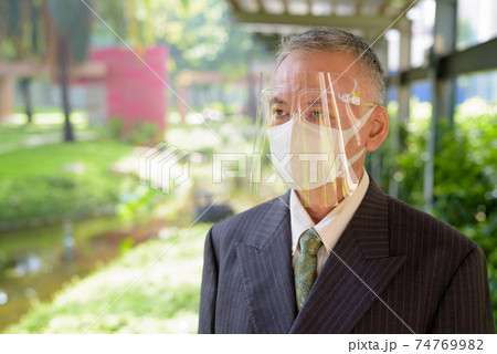 Mature Japanese businessman with mask and face shield thinking at the park 74769982