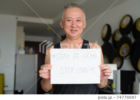 Happy mature Japanese man holding Stay At Home sign while under quarantine due to covid-19 pandemic 74770097