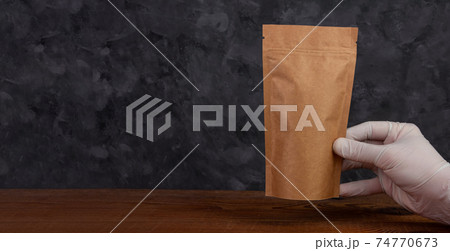 Pouch bag mockup banner. Hand in protective gloves holds brown kraft paper pack with coffee beans wooden background. 74770673