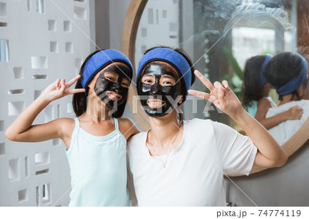 Smiling beautiful little girl and her mother in peaceful hand gestures using black clay face masks 74774119