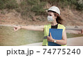 Young female construction worker with white helmet and face mask pointing with her hand at construction site. 74775240