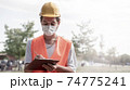 Young male construction worker with yellow helmet and face mask taking notes at construction site. New normality after covid-19 outbreak. 74775241