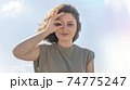 Charming young woman doing ok gesture with her hand and looking through OK sign smiling with happy face against sky background. Facial expression. 74775247