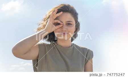 Charming young woman doing ok gesture with her hand and looking through OK sign smiling with happy face against sky background. Facial expression. Charming young woman doing ok gesture with her hand and looking through OK sign smiling with happy face against sky background. Facial expression. 74775247