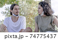 Two young friends with face masks sitting on the green grass smiling and having fun. Meeting with friends in the park during the coronavirus epidemic. 74775437